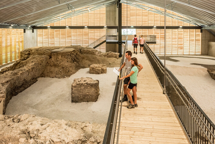 Römerburgus Oberranna in Engelhartszell: In dieser Halle mit hoher Decke und Holzwänden befinden sich einige Menschen, die sich auf einem Laufsteg zwischen erhöhten Erdformationen bewegen. Die Umgebung scheint einem archäologischen oder historischen Ausstellungsraum zu ähneln, in dem die Besucher einen Einblick in vergangene Zeiten erhalten können.