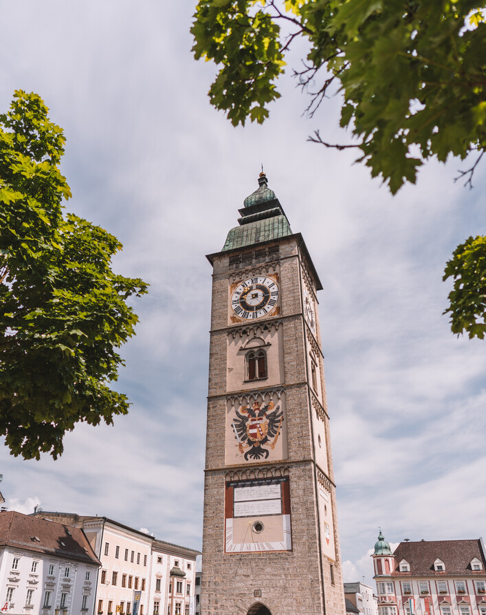 Stadtturm in Enns: Ein hoher Turm mit einer kunstvoll verzierten Fassade thront inmitten eines malerischen Stadtbilds. Die Uhr am Turm ist deutlich erkennbar, darüber befindet sich ein ornamentierter Aufbau. Grüne Baumkronen rahmen den Turm und verleihen dem Gesamtbild eine harmonische Atmosphäre.
