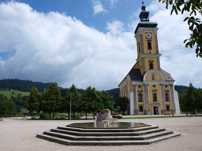 Stiftskirche Waldhausen im Strudengau in Waldhausen im Strudengau: Ein Kirchenbau mit einem hohen Turm, in gelblicher Fassade gehalten, umgeben von Bäumen und einem Brunnen auf einem Platz davor. Das Gebäude weist eine traditionelle Architektur auf und wirkt einladend.