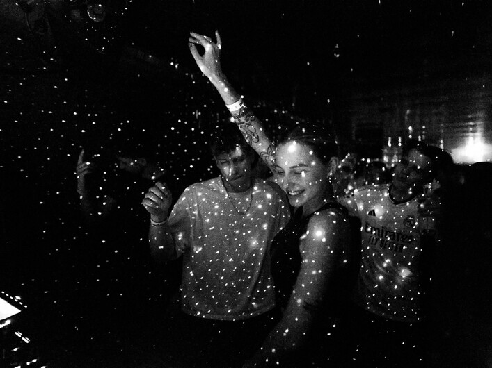 Schwarz-Weiß-Aufnahme von Menschen, die in einem dunklen Raum tanzen, umgeben von Lichtpunkten, die wie Sterne im Raum wirken