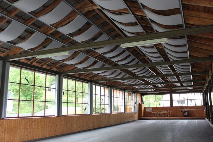 Asphaltbahnen Aubach in St. Martin im Mühlkreis: Ein großer Innenraum mit hoher Holzdecke, die an ein Wellendach erinnert. Große Fenster an den Wänden lassen viel Tageslicht herein. Der Boden ist mit Dielen belegt, und es sind einige Sitzmöbel zu sehen.