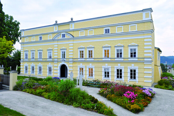 Schloss Mattighofen in Mattighofen: Ein großes, gelb gestrichenes Gebäude mit einer aufwendigen Fassade, umgeben von üppigen Blumenbeeten und grünen Rasenflächen. Das Gebäude hat mehrere Stockwerke und Balkone sowie weiße Säulen und dekorative Elemente an der Fassade. Die Umgebung wirkt gepflegt und lädt zum Verweilen ein.