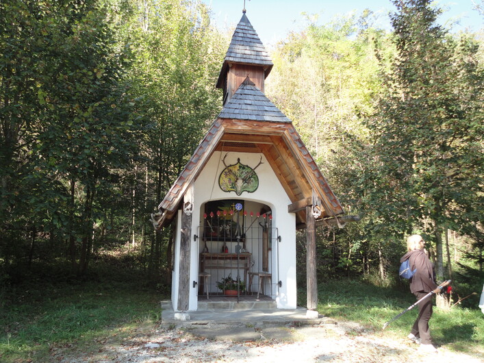 Hubertuskapelle in Seewalchen am Attersee: Das Bild zeigt ein kleines, traditionelles Gebäude mit einem spitzen Dach und Holzelementen. Es befindet sich inmitten eines waldigen Gebiets mit grüner Vegetation. Vor dem Gebäude ist eine Person zu sehen, die den Weg entlanggeht.
