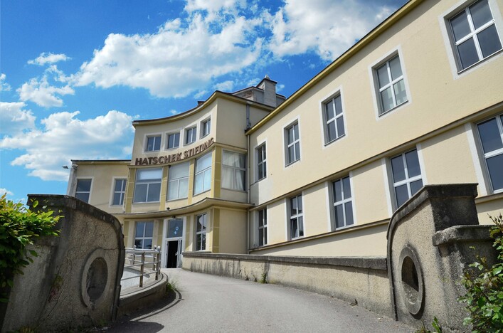 OKH - Offenes Kulturhaus Vöcklabruck in Vöcklabruck: Ein mehrstöckiges Gebäude mit einer hellen Fassade und geschwungenen Dächern. Der Eingang ist durch Treppen und Säulen gekennzeichnet. Davor erstreckt sich ein gepflasterter Vorplatz mit üppigem Grün.