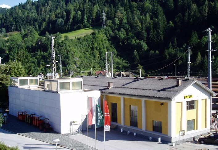 Ein modernes Gebäude mit einer gelben Fassade und mehreren Antennen steht in einer grünen Berglandschaft. Es gibt auch einige Flaggen und orangefarbene Behälter im Vordergrund.