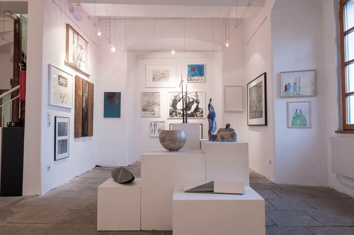 Galerie Schloss Puchheim in Attnang-Puchheim: Eine Galerie-Ausstellung mit verschiedenen Kunstwerken wie Gemälde, Skulpturen und Zeichnungen an den Wänden und auf Podesten. Die Werke zeigen eine Vielfalt von Stilen und Themen. Der Raum ist hell und geräumig, mit Fenstern, die natürliches Licht einfallen lassen. Es herrscht eine ruhige, kunstvolle Atmosphäre.