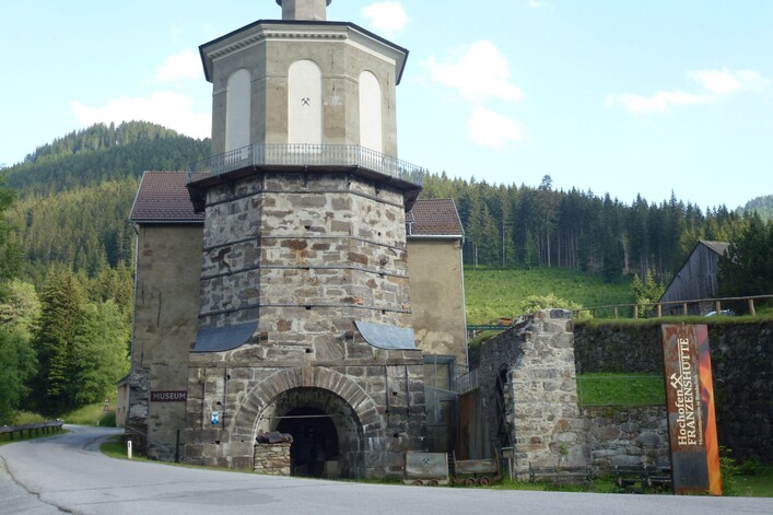 Ein historisches Gebäude mit einem runden Turm und einer steinernen Fassade. Umgeben von grünen Bergen und einer ruhigen Straße.