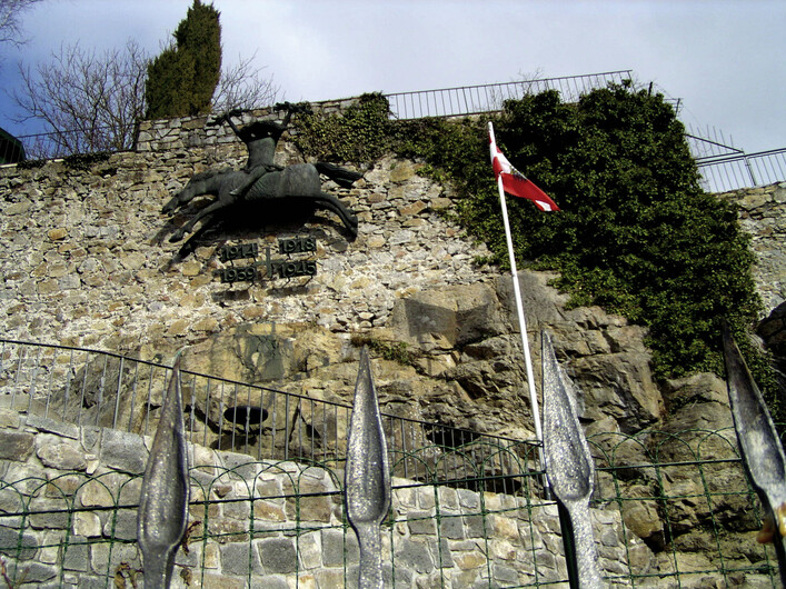 Kriegsmahnmal in Schärding: Eine Steinmauer mit einem eisernen Geländer ist zu sehen. Darauf befindet sich eine Bronzeskulptur eines Tieres. Eine Flagge ist an der Mauer angebracht. Die Umgebung ist von Bäumen und Sträuchern bewachsen.