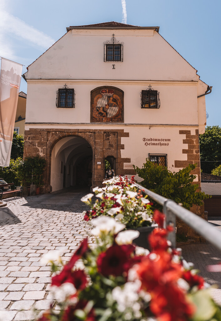 Stadtmuseum Schärding in Schärding: Ein zweistöckiges Gebäude mit traditioneller Architektur. Das Erdgeschoss ist mit einem schmuckvollen Torbogen verziert, über dem ein Fresko eine Heiligenfigur zeigt. Blumengirlanden säumen den Eingang. Die Fassade ist hell verputzt und durch Fenster, Verzierungen und ein Ziegeldach strukturiert. Insgesamt vermittelt das Gebäude einen historischen und malerischen Eindruck.