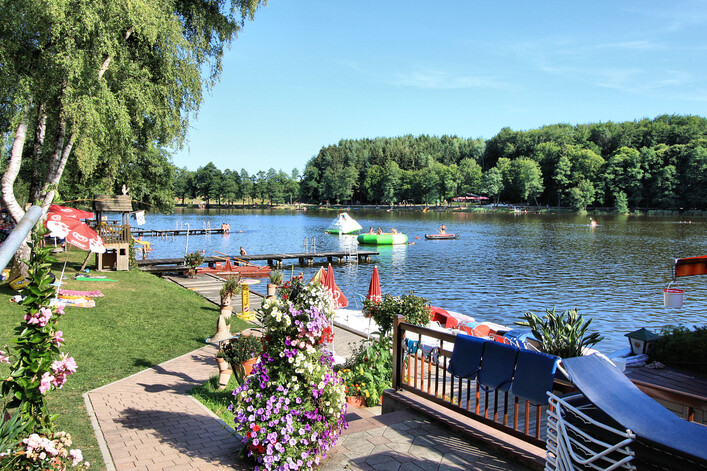 Strandbad Holzöstersee in Franking: Ein idyllischer See, umgeben von grünen Bäumen und Sträuchern. Es gibt einen hölzernen Steg mit mehreren Booten und aufblasbaren Spielgeräten im Wasser. Entlang des Ufers sind Tische und Sonnenschirme aufgestellt, die eine einladende Atmosphäre schaffen.