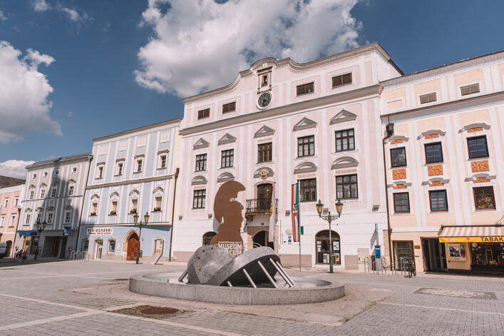 Museum Lauriacum in Enns: Ein großes, mehrstöckiges Gebäude mit einer beeindruckenden Fassade. Die Architektur ist im traditionellen Stil mit vielen Verzierungen und Ornamenten gestaltet. Vor dem Gebäude befindet sich ein Brunnen mit einer Statue. Insgesamt vermittelt das Bild einen historischen, eleganten Eindruck.