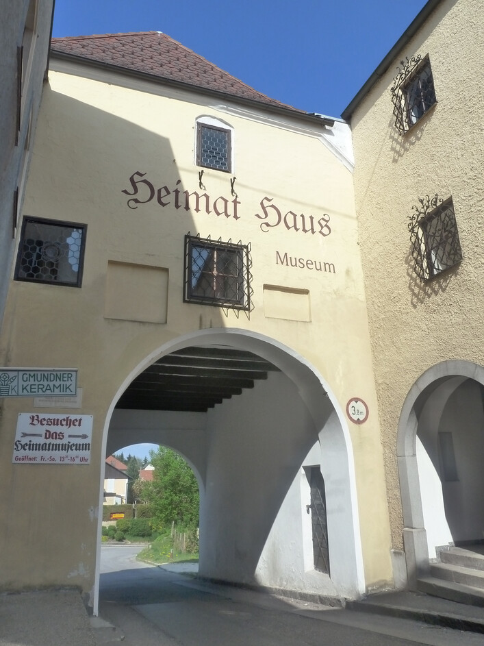 Heimatmuseum Obernberg am Inn in Obernberg am Inn: Auf dem Bild ist ein historisch anmutendes Museumsgebäude mit einem Torbogen zu sehen. Es trägt den Namen "Heimat Haus Museum" in deutscher Schrift. Das Gebäude hat ein traditionelles Aussehen mit Natursteinbogen und Fenstern. Außerdem ist ein Schild für Öffnungszeiten und Eintrittspreise zu erkennen.
