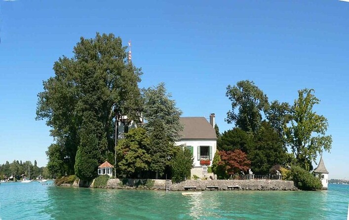 Schloss Litzlberg in Seewalchen am Attersee: Das Bild zeigt ein malerisches Haus, das sich auf einer Insel inmitten eines ruhigen Sees befindet. Das Gebäude ist von üppiger Vegetation umgeben und scheint ein traditionelles Landhaus-Styling zu haben. Im Hintergrund sind weitere Bäume und eine klare Himmelskulisse zu sehen.