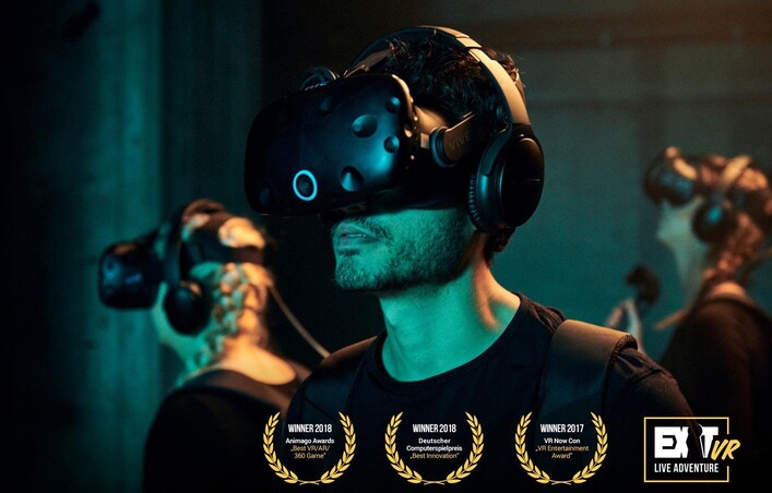 Eine Person mit einer Virtual-Reality-Brille und Kopfhörern erlebt ein immersives Abenteuer. Im Hintergrund sind weitere Teilnehmer zu sehen, die ebenfalls VR-Technologie nutzen.