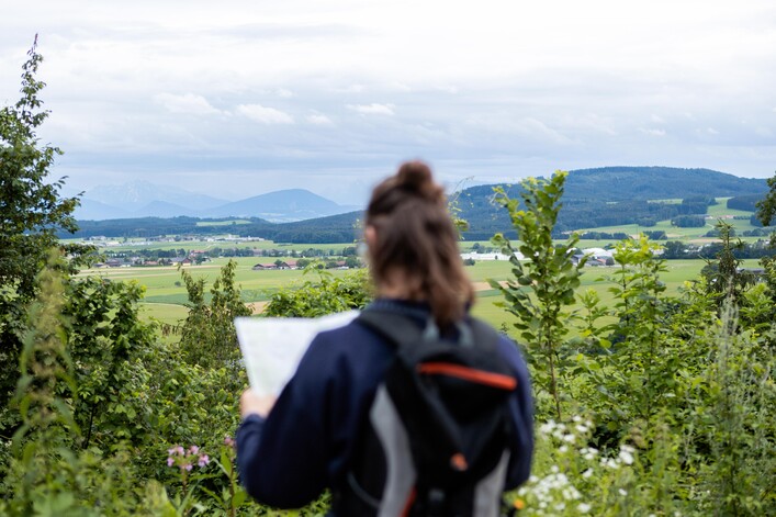 Schlossberg Friedburg mit Weinberg in Lengau: Eine Person, die eine Jacke trägt, steht in einer grünen Umgebung mit Bergen im Hintergrund. Die Landschaft ist von Natur umgeben, und die Person scheint eine malerische Aussicht zu genießen.