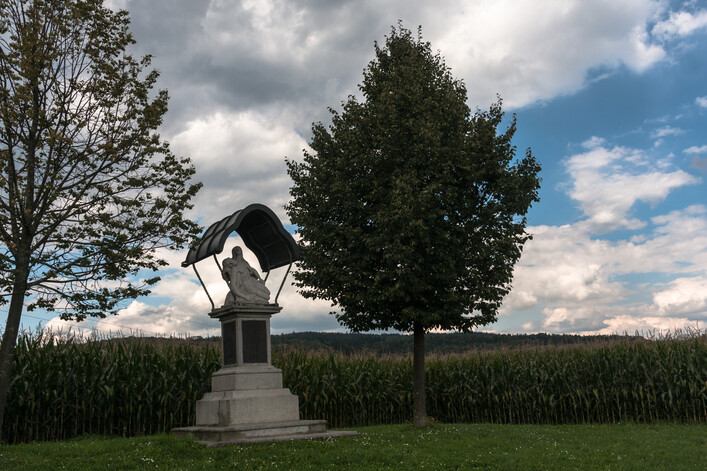 Das Steinerne Kreuz in Rufling in Leonding: Eine Statue eines Mannes in einer Nische, umgeben von Bäumen auf einer Wiese. Im Hintergrund ist ein bewölkter Himmel zu sehen.