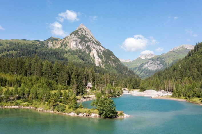 Eine malerische Landschaft mit einem klaren blauen See und grünen Bergen im Hintergrund. Die Sonne scheint und der Himmel ist teilweise bewölkt.