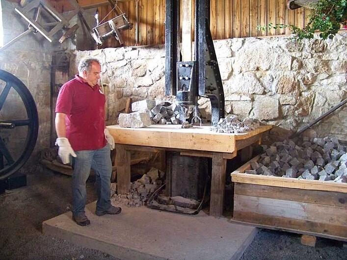 Granitlehrpfad Erlebniswelt Granit und Arena Granit in St. Martin im Mühlkreis: Eine Person in einem roten Hemd steht in einer Werkstatt mit einer Holzbank, auf der verschiedene Werkzeuge und Gesteinsbrocken liegen. Die Wände sind aus Stein, und an der Decke hängen weitere Werkzeuge. Rechts sind Regale aufgestellt, auf denen weitere Steine und Materialien gelagert sind.