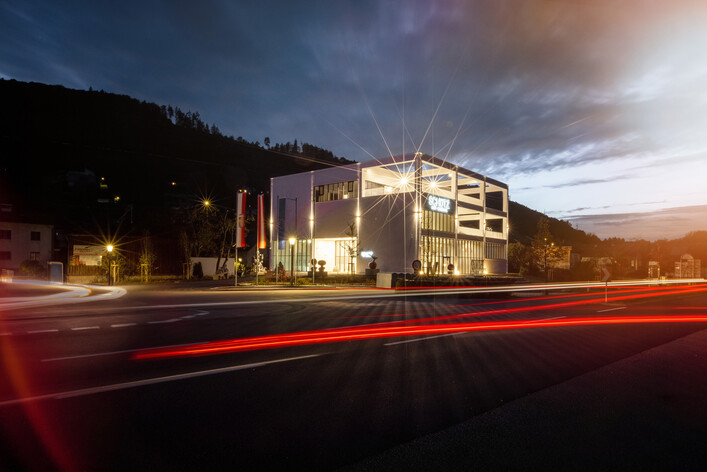 Schütz Art Museum in Engelhartszell: Ein modernes Gebäude in einer hügeligen Umgebung, beleuchtet in der Abenddämmerung. Verschiedene Lichtquellen, wie Straßenlaternen und Scheinwerfer, erzeugen ein stimmungsvolles Spiel von Helligkeit und Schatten. Die architektonische Gestaltung des Gebäudes zeigt klare Linien und Formen.
