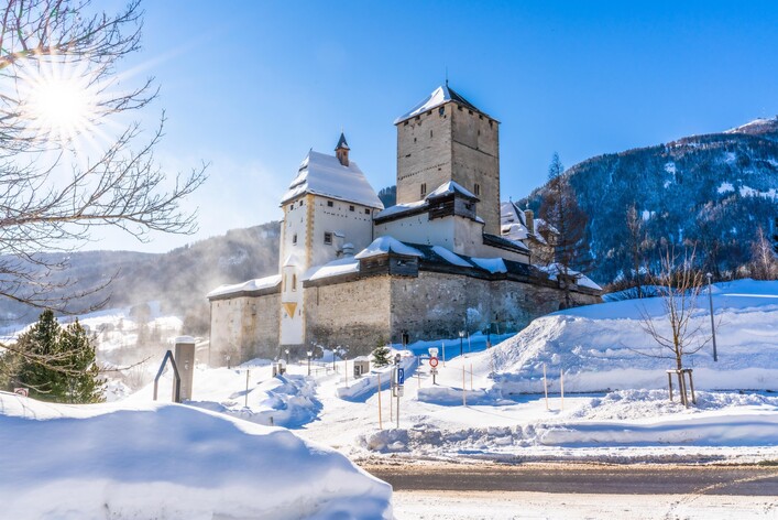 Eine alte Burg in schneebedeckter Landschaft. Der Himmel ist klar und die Sonne strahlt.