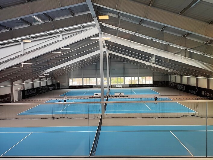 Tennis-Zentrum Danubis in Mauthausen: Eine große, überdachte Sportanlage mit mehreren Tennisplätzen. Im Inneren gibt es ein geräumiges Hallengebäude mit hoher Decke, das die Tennisplätze beherbergt. Die Böden der Plätze sind in blau- und hellgrauen Farbtönen gehalten. Insgesamt bietet die Anlage Möglichkeiten für sportliche Betätigung in einer modernen und zweckmäßigen Umgebung.
