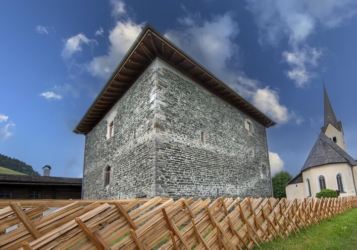 Ein historisches Gebäude aus Stein mit einem Holzdach. Im Hintergrund ist ein Kirchturm zu sehen unter einem teilweise bewölkten Himmel.