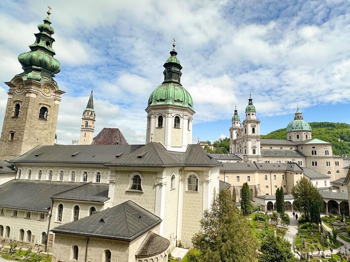 Eine beeindruckende Klosteranlage mit vielen Türmen und grünen Dächern. Der Himmel ist teilweise bewölkt und es sind verschiedenen Pflanzen im Vordergrund zu sehen.
