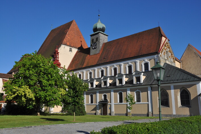 Stiftskirche Baumgartenberg in Baumgartenberg: Ein historisches Gebäude mit charakteristischer Architektur und Türmen. Das Hauptgebäude hat eine helle Fassade mit Bögen und Elementen im Stil alter Baukunst. Aufragende Dächer und Türme mit spitzen Dächern prägen die Silhouette. Umgeben ist das Ensemble von üppiger Vegetation.