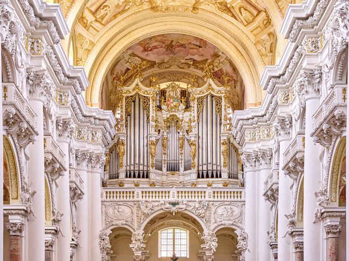 Brucknerorgel in St. Florian: Eine prächtige Kirche mit reich verzierter Fassade und Orgelprospekt. Die Architektur ist im prunkvollen Barockstil gehalten, mit hohen Bögen, Säulen und einer üppigen Ornamentik. Der Raum ist hell erleuchtet und strahlt eine feierliche Atmosphäre aus.