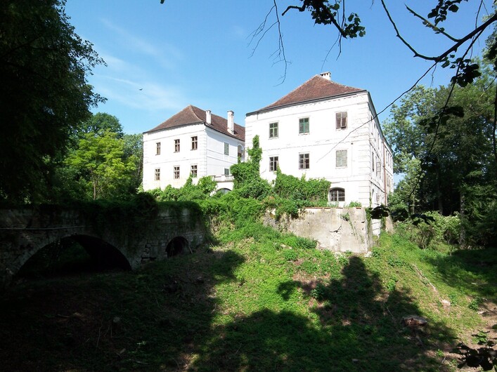 Schloss Katzenberg in Kirchdorf am Inn: Ein geschichtsträchtiges Gebäude mit traditioneller Architektur, eingebettet in eine üppige grüne Umgebung. Die Fassade und die Dächer zeugen von einem älteren Baustil. Eine Brücke über einen kleinen Fluss oder Bach führt zum Hauptgebäude.