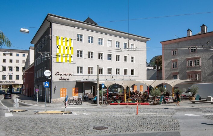 Ein modernes Gebäude mit auffälliger Beschriftung und Außenbereich für Gäste. Die Straße ist belebt und zeigt eine Mischung aus modernen und historischen Architektur.