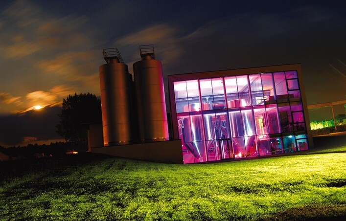 Ein modernes Gebäude mit großen Fenstern, das in bunten Neonfarben beleuchtet ist. Im Hintergrund stehen zwei silberne Silos auf einer grünen Wiese.