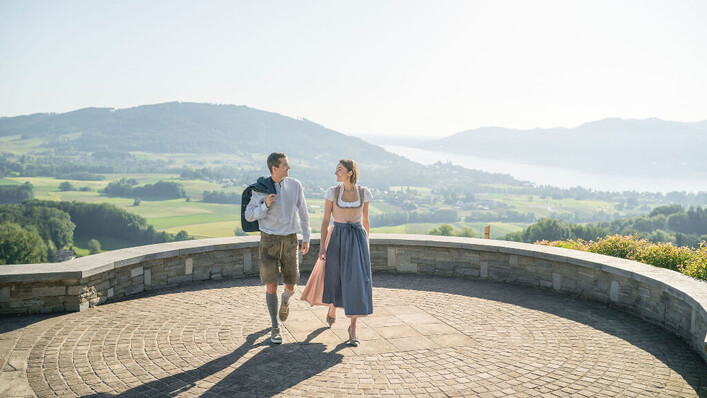 Frau und Mann auf einem Aussichtsplatz bei der Kronbergkapelle mit Hügeln und dem Attersee im Hintergrund.
