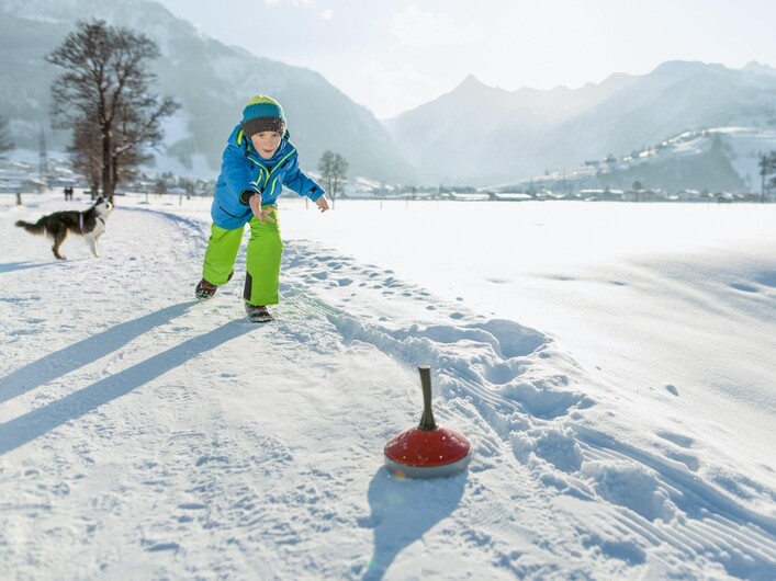 Ein Junge spielt im Schnee und versucht, eine rote Kelle zu treffen. Im Hintergrund sind Berge und ein Hund zu sehen.