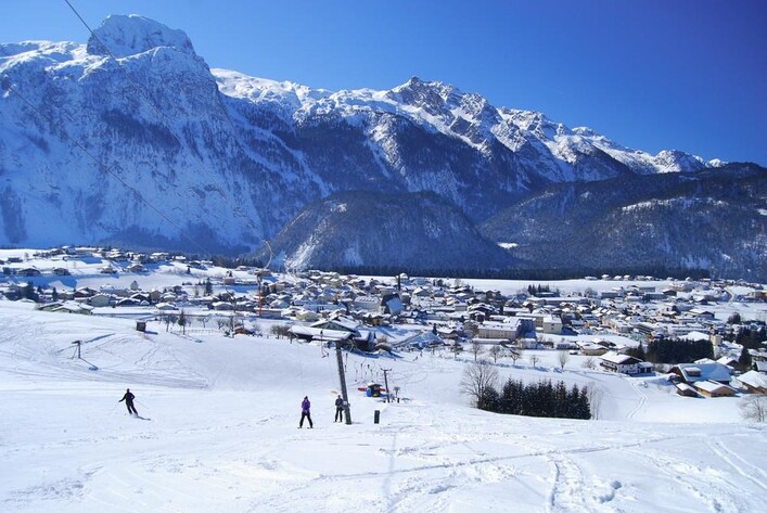 Eine schneebedeckte Landschaft mit Bergen im Hintergrund und einem kleinen Dorf. Skifahrer sind auf der Piste zu sehen, unter einem klaren blauen Himmel.