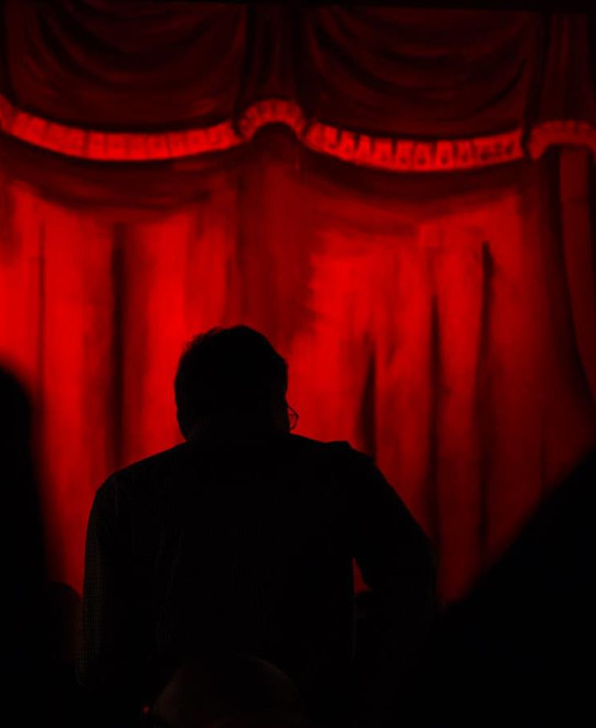 Kellerbühne Puchheim in Attnang-Puchheim: Vor einem dunkelroten, samtigen Bühnenhintergrund ist eine schwarz silhouettierte Menschengestalt zu erkennen. Die Figur steht im Mittelpunkt des Bildes und ist von der Rückseite zu sehen. Die Atmosphäre erscheint düster und geheimnisvoll.