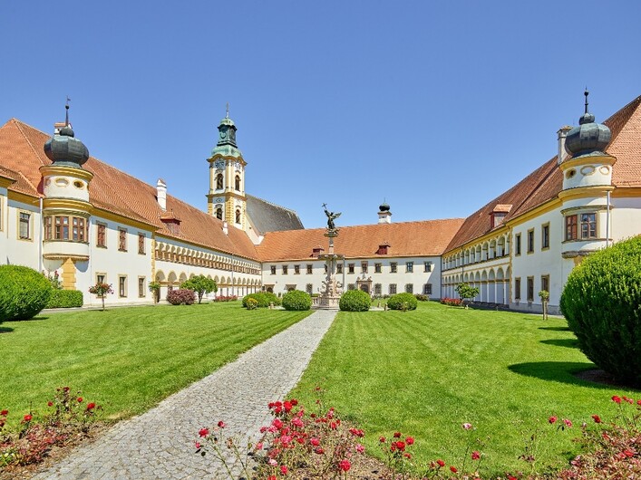 Stift Reichersberg in Reichersberg: Ein historisches Gebäude mit traditioneller Architektur und Türmen in einem gepflegten Garten. Die Fassade ist mit Ornamenten und Dekorationen verziert, umgeben von Grünflächen und Blumenbeeten. Ein geräumiger Innenhof mit einem Springbrunnen bildet das Zentrum der Anlage.