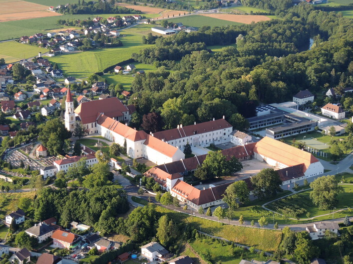 Schloss / Stift Ranshofen in Braunau am Inn: Ein historisches Gebäude mit traditioneller Architektur thront inmitten einer hügeligen, grünen Landschaft. Mehrere Türme und Dächer sind zu sehen, die auf ein großes Areal hinweisen. Umliegende Wohnhäuser und Wälder umgeben das Hauptgebäude, das den Mittelpunkt der Ansicht bildet.