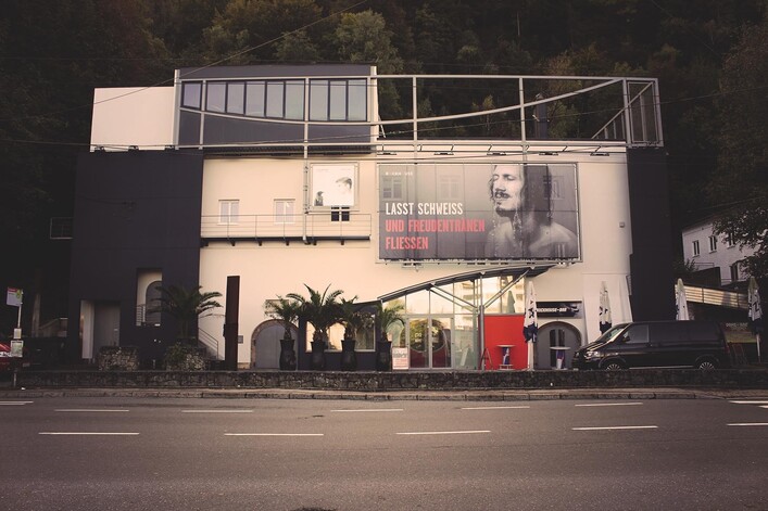 Ein modernes Gebäude in Blau mit einer großen Werbetafel für "Rockhouse". Vor dem Gebäude stehen mehrere Autos und Palmen.