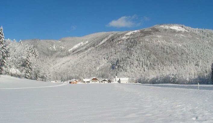 Eine verschneite Landschaft mit einem klaren blauen Himmel. Im Hintergrund sind bewaldete Hügel und einige Gebäude sichtbar.