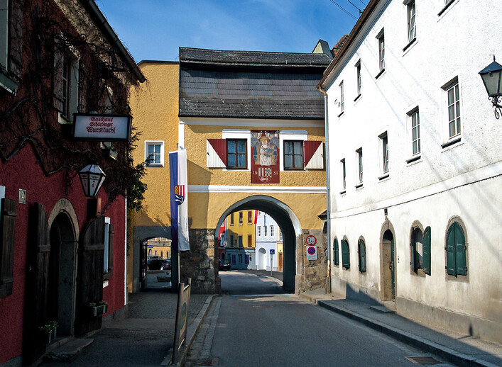 Passauer Tor in Schärding: Eine historische Gebäudefassade mit traditioneller Architektur, die einen Durchgang mit einem Bogentorweg bildet. Mehrere farbenfrohe Gebäude säumen die Straße, die von Bäumen und Grünflächen umgeben ist.