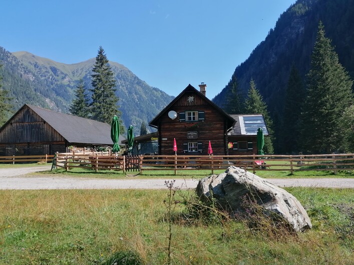 Ein rustikales Holzhaus inmitten einer grünen Wiese, umgeben von hohen Bergen. Im Vordergrund steht ein großer Stein und die Aussicht ist klar und sonnig.