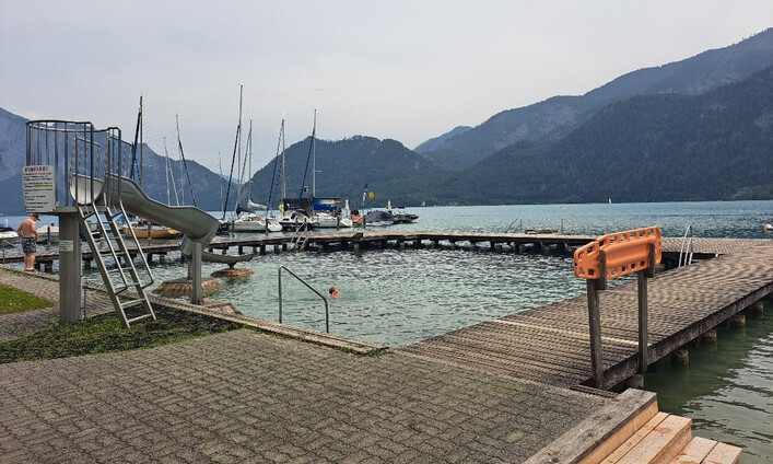 Strandbad Unterach in Unterach am Attersee: Das Bild zeigt einen Bootsanleger mit Blick auf einen See, umgeben von hohen Bergen. Am Steg befinden sich mehrere Boote und eine Rutsche ins Wasser.