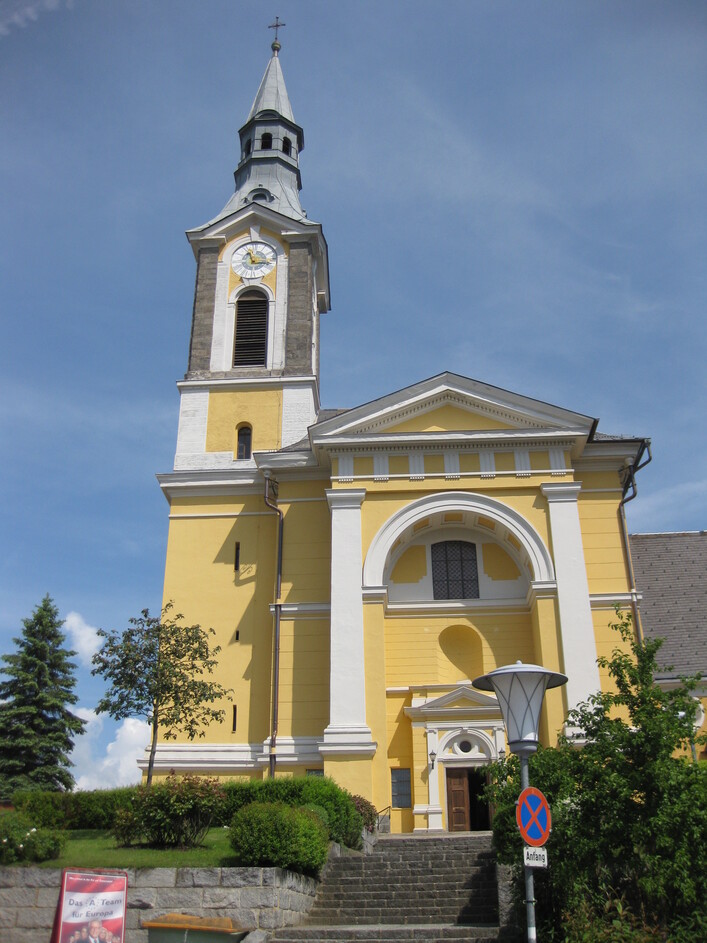 Mühlviertler Dom in Niederkappel: Eine große, gelblich gestrichene Kirche mit einem hohen Glockenturm und einer Uhr an der Fassade. Vor dem Haupteingang der Kirche führen Stufen hinauf und es gibt einen kleinen Garten mit Bäumen. Insgesamt zeigt das Bild ein traditionelles, historisches Kirchengebäude in einer ländlichen Umgebung.