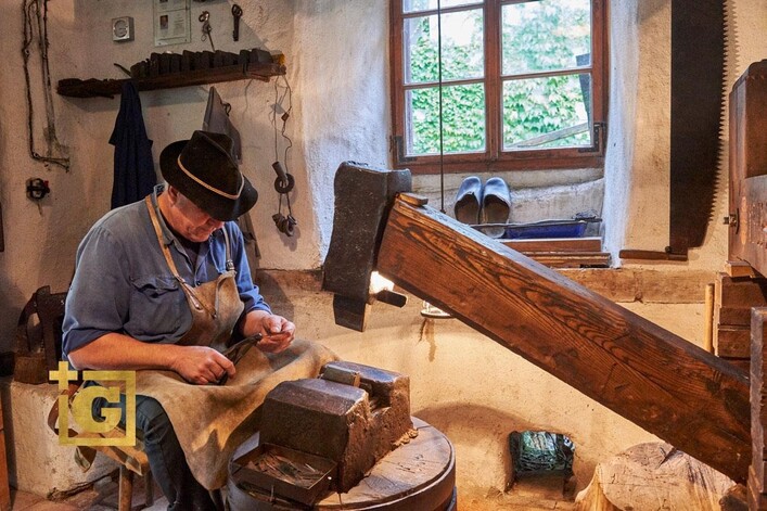 Ein Handwerker arbeitet konzentriert in einer traditionellen Werkstatt. Im Hintergrund sind Werkzeuge und eine Fensteransicht zu sehen.