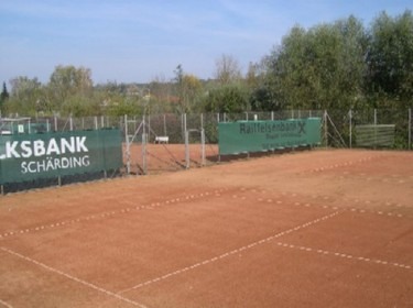 Tennisplatz Union Schärding in Schärding: Eine Tennisanlage mit mehreren Tennisplätzen umgeben von Bäumen und Sträuchern. Im Vordergrund ist ein grüner Tennisplatz mit einem Netz zu sehen. Hinter dem Platz befinden sich weitere Tennisplätze und eine Werbemarkise.