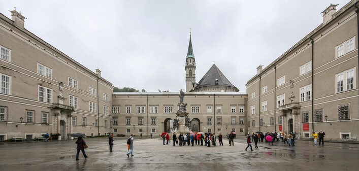 Domplatz (Cathedral Square) in the historic centre of the city of Salzburg, as seen from the cathedral. The square is surrounded by a part of Salzburg Residence. In the center: Wallistrakt which houses lecture halls of the University of Salzburg. In the background: Franciscan church.