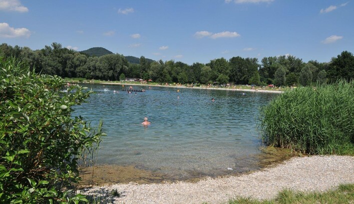 Ein ruhiger See umgeben von Bäumen und Gras. Menschen genießen das Wasser an einem sonnigen Tag.