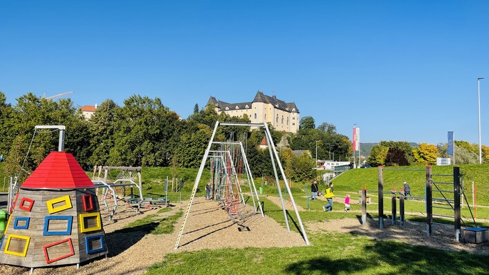 Generationen-Motorikpark® in Grein: Ein großer Spielplatz mit bunten Spielgeräten, Schaukeln und Klettergerüsten. Im Hintergrund ist ein malerisches, historisches Gebäude mit Türmen zu sehen, umgeben von grünen Bäumen. Kinder spielen fröhlich auf dem Spielplatz.