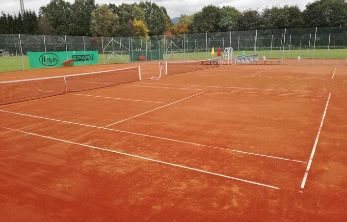 Ein Tennisplatz aus roter Erde mit klaren Linien. Im Hintergrund sind Bäume und Tennisstühle zu sehen.